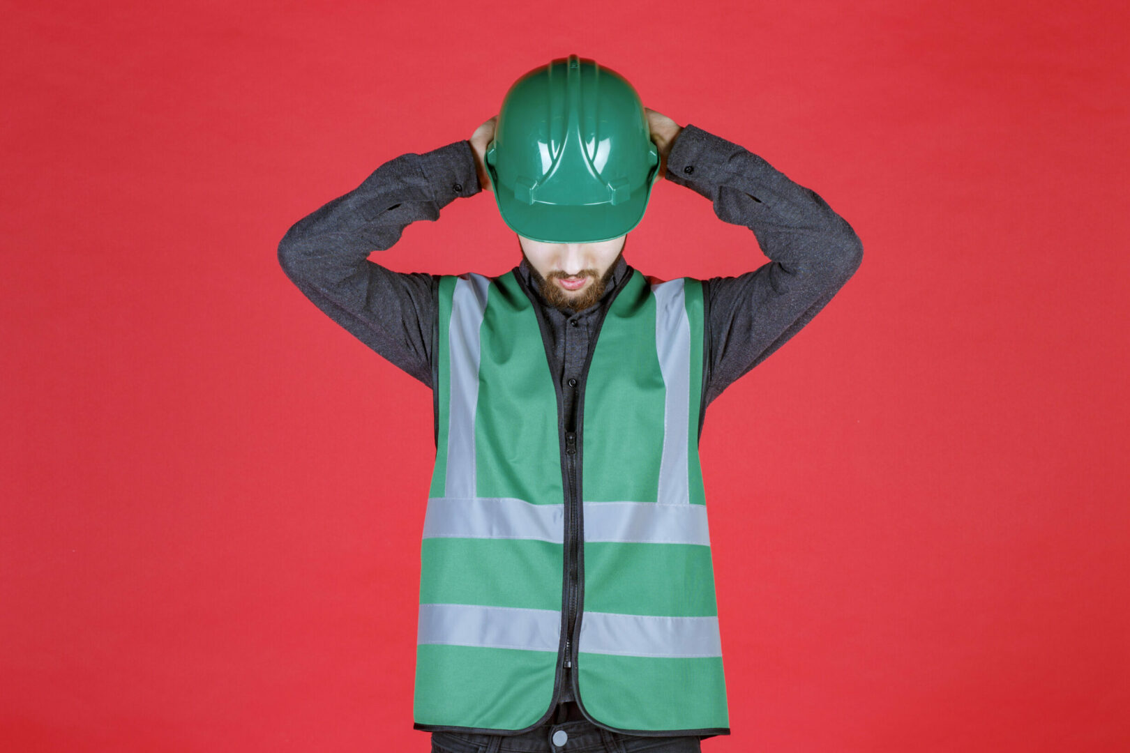 Fond rouge. C'est un ingenieur avec un uniforme et un casque vert. Il joint ses mains derrière la tête, sur son casque, la tête penchée en avant.