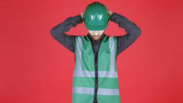 Fond rouge. C'est un ingenieur avec un uniforme et un casque vert. Il joint ses mains derrière la tête, sur son casque, la tête penchée en avant.