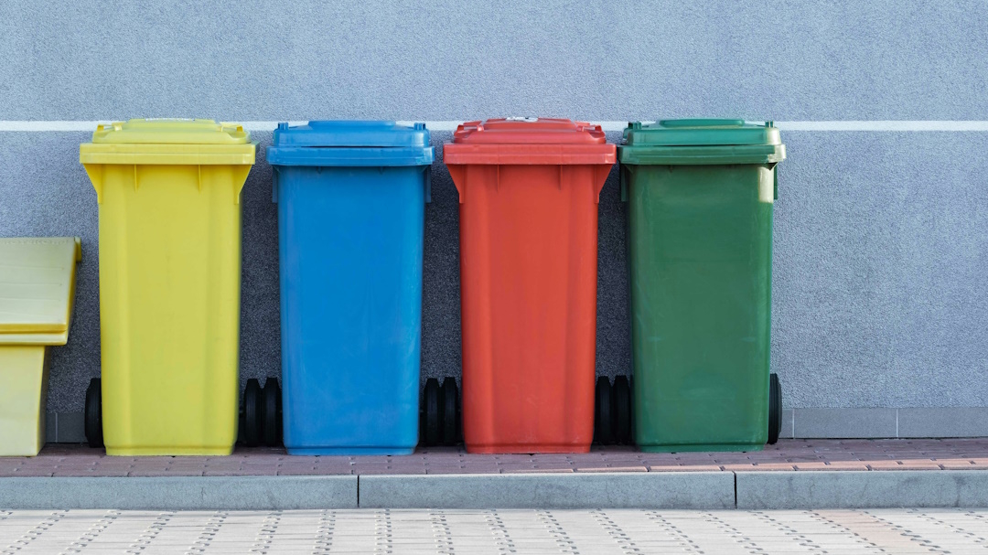 Photo dans la rue de 4 poubelles, chacune une couleur différente (jaune, bleu, rouge, vert). Elle sont sur le trottoir devant un mur bleu ciel uni. Ces différentes couleurs de poubelles sont une aide précieuse pour gérer les déchets et améliorer le suivi des bennes.