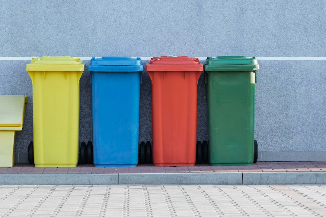 Photo dans la rue de 4 poubelles, chacune une couleur différente (jaune, bleu, rouge, vert). Elle sont sur le trottoir devant un mur bleu ciel uni. Ces différentes couleurs de poubelles sont une aide précieuse pour gérer les déchets et améliorer le suivi des bennes.